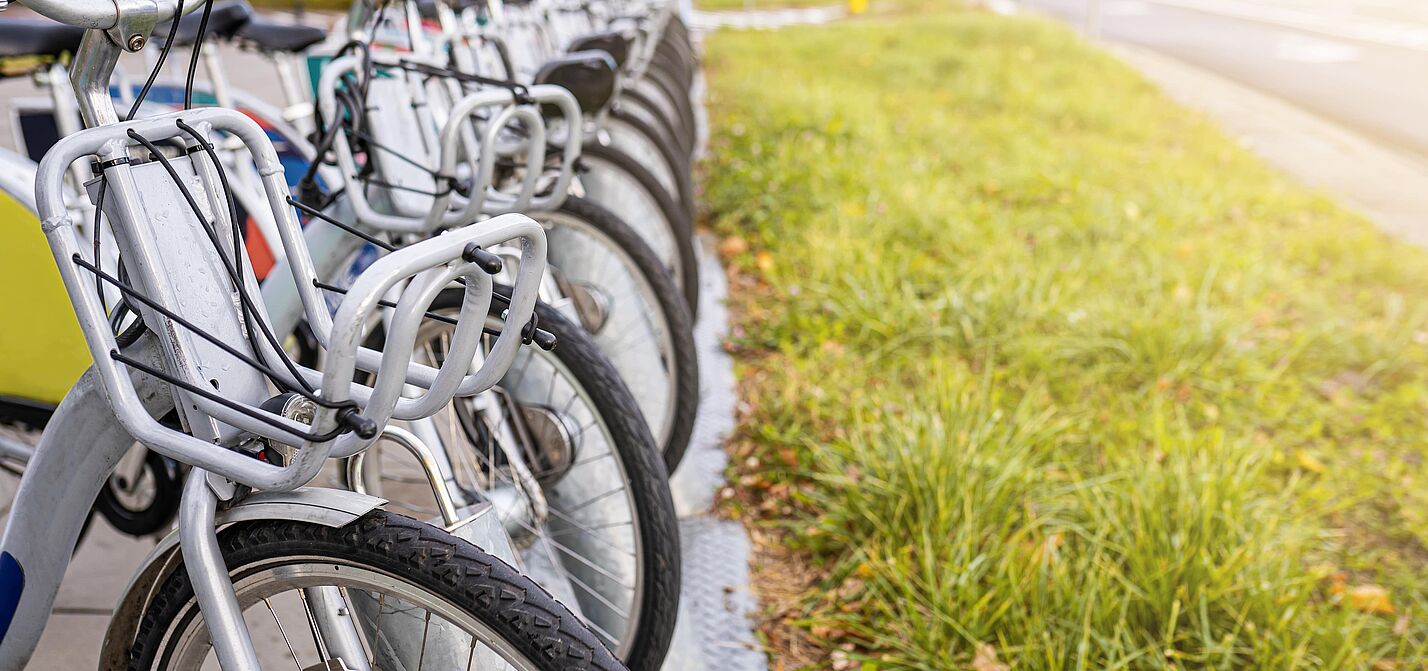 Öffentliche Leihfahrräder an einer Bike-Sharing-Station Eine Reihe von öffentlichen Leihfahrrädern mit silbernen Rahmen und grauen Körben am Lenker steht ordentlich nebeneinander an einer stationsgebundenen Verleihstation. Die Fahrräder sind auf einem gepflasterten Gehweg abgestellt, neben dem eine Grünfläche verläuft.